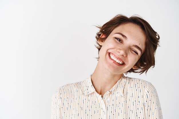 Retrato sincero de close-up de uma linda mulher inclinando a cabeça e sorrindo feliz expressando alegria e positividade, em pé na parede branca