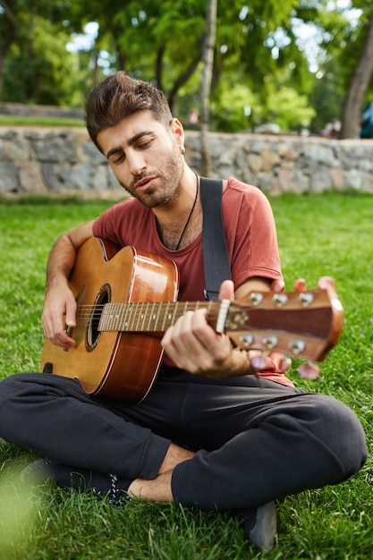 Retrato vertical ao ar livre de cara bonito hipster sentado na grama do parque tocando violão