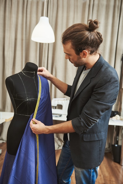 Retrato vertical de adulto bonito caucasiano masculino estilista com penteado elegante com roupa elegante em seu estúdio, trabalhando no vestido novo para a coleção de roupas de inverno