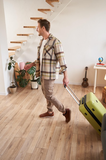 Retrato vertical de um jovem bonito caminhando em casa com uma mala indo de férias deixando sua