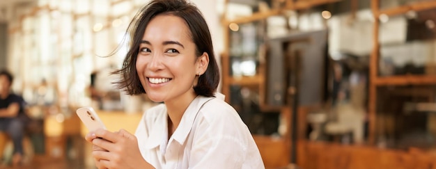 Foto grátis retrato vertical de uma elegante mulher asiática sentada em um café bebendo café e usando um smartphone