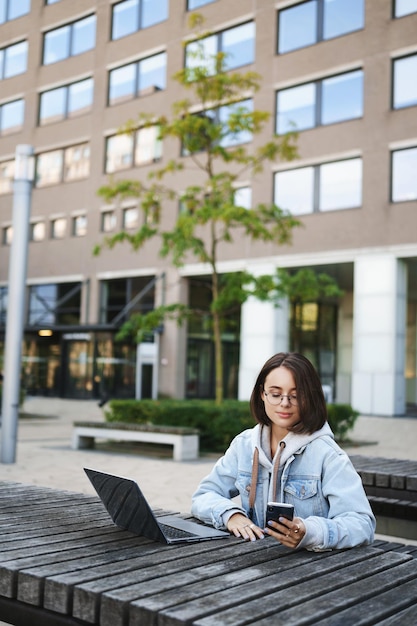 Retrato vertical de uma jovem aluna bonita sentada do lado de fora com o laptop olhando para a tela do celular enquanto trabalhava smm respondendo às perguntas dos clientes estudando on-line