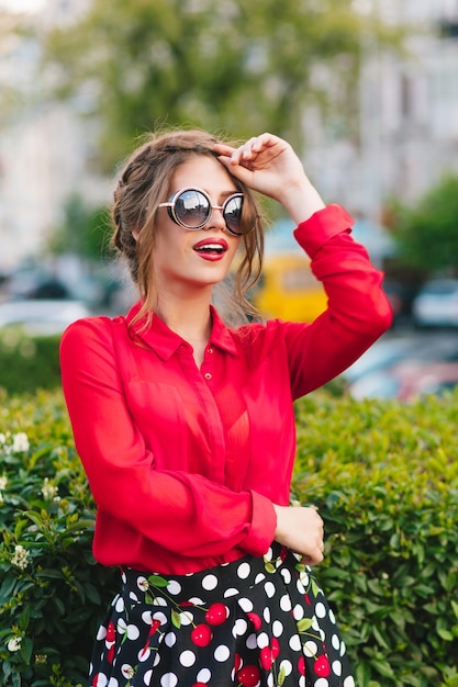 Retrato vertical de uma menina bonita em óculos de sol, posando para a câmera no parque. Ela usa blusa vermelha, saia preta e penteado bonito. Ela está olhando para longe.