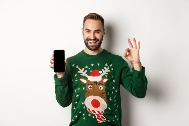 Reveillon, feriados e comemorações. Comprador do sexo masculino jovem satisfeito mostrando o sinal de OK e a tela do celular, recomendando um aplicativo, em pé sobre um fundo branco.