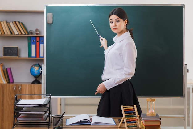 rigorosa jovem professora em pé na frente do quadro-negro e aponta para o quadro-negro com ponteiro na sala de aula