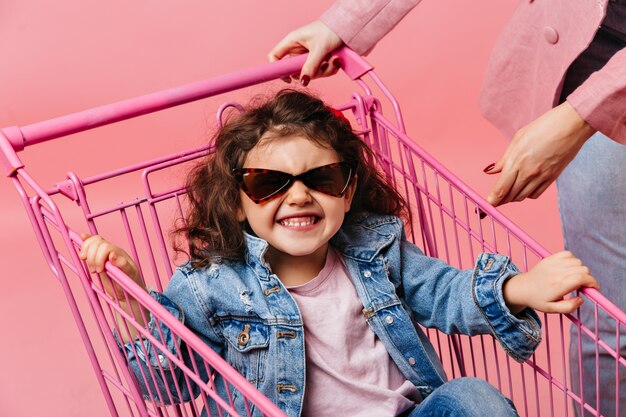 Rindo criança pré-adolescente sentada no carrinho de compras. Foto de estúdio de criança encaracolada em jaqueta jeans e óculos escuros.