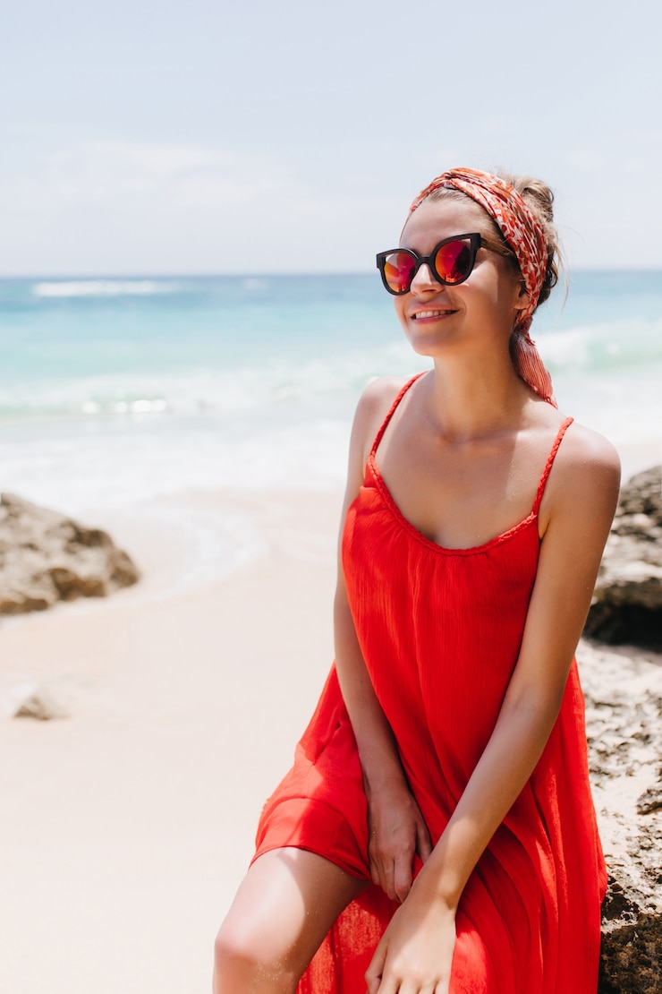 Foto grátis rindo mulher branca bronzeada sentada na praia. retrato ao ar livre do inspirado modelo feminino caucasiano em vestido vermelho, passando os momentos de lazer perto do mar.