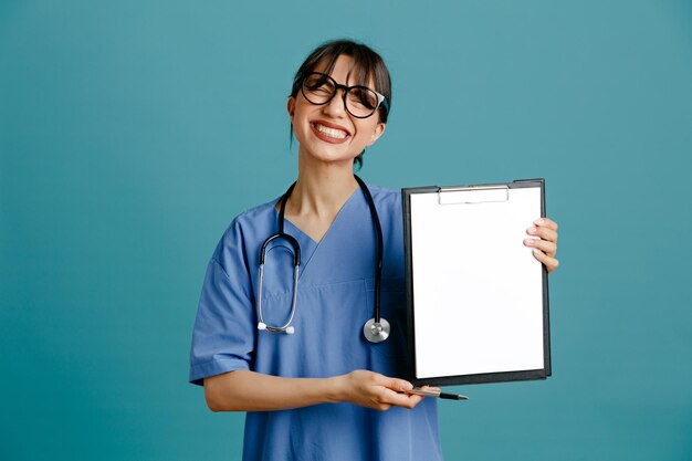 Rindo segurando a prancheta jovem médica usando estetoscópio uniforme de quinto isolado em fundo azul