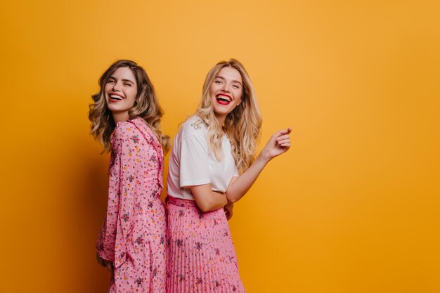 Rindo senhoras da moda posando juntas durante a sessão de fotos interna Adoráveis irmãs adultas em pé no fundo amarelo