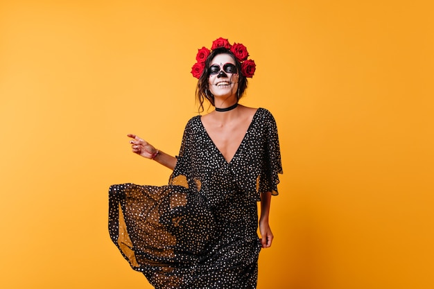 Rindo zumbi feminino com rosas no cabelo dançando no estúdio. garota feliz com maquiagem mexicana comemorando o dia das bruxas.