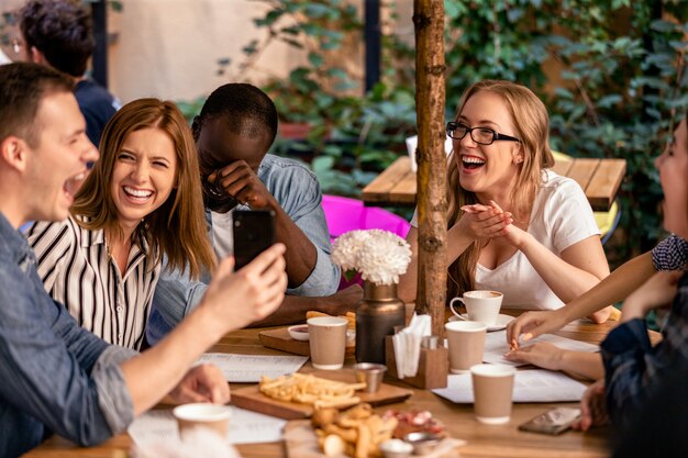 Riso sincero e mostrando imagens no smartphone na reunião casual com os melhores amigos no terraço do restaurante