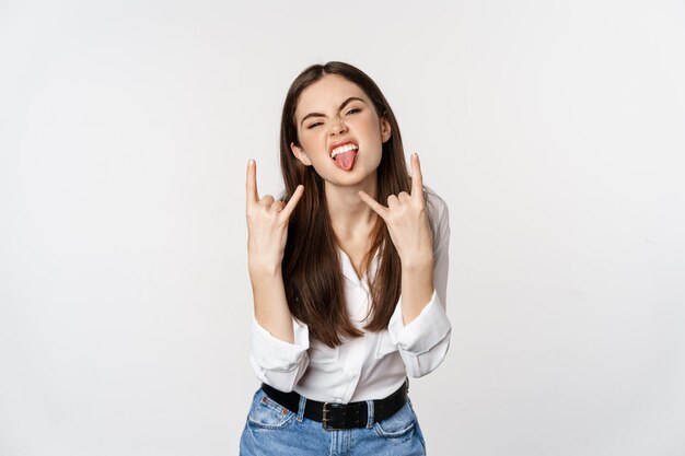 Rock n roll. Jovem mulher mostrando rock, sinal de heavy metal, se divertindo, em pé despreocupado contra um fundo branco.