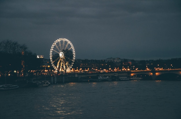 Roda-gigante rodeada por um rio e edifícios sob um céu nublado durante a noite em Paris