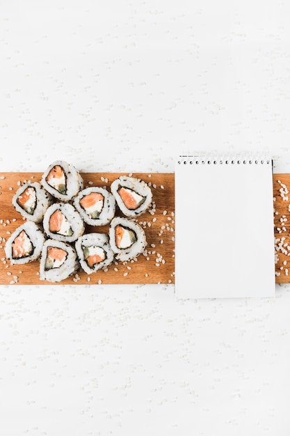 Rolos de sushi e bloco de notas em espiral na bandeja de madeira com salpicos de arroz cru em pano de fundo branco
