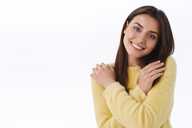 Foto grátis romântica e atraente fêmea de suéter amarelo, abraçando-se, enrole o corpo com as próprias mãos, sentindo-se confortável e feliz, sorrindo, alegremente, o amor está no ar, romance e conceito de beleza