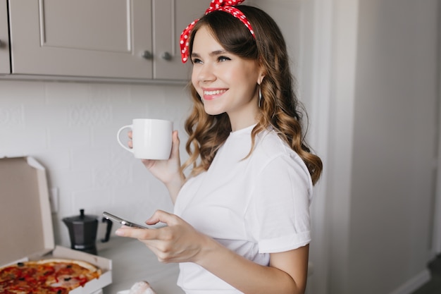 Romântica garota caucasiana com cabelos ondulados em pé na cozinha perto de pizza. mulher branca relaxada, desfrutando de chá com um sorriso.