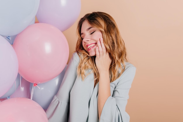 Romântica garota encaracolada em traje azul, posando com os olhos fechados e sorriso sincero. foto interna da despreocupada senhora branca comemorando aniversário com balões.