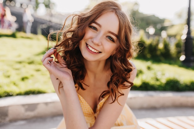 Romântica garota ruiva com olhos claros, sorrindo para a natureza. Mulher jovem e sonhadora em roupas amarelas, brincando com seu cabelo ruivo cacheado.