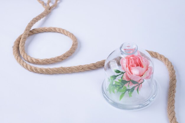 Rosa em copo de vidro com corda na superfície branca.