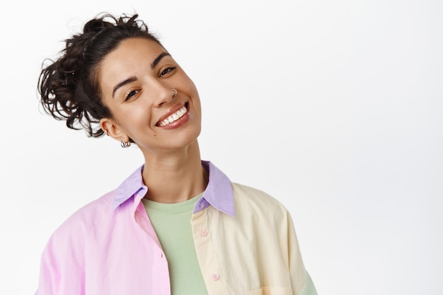 Foto grátis rosto de menina morena feliz com cabelo penteado encaracolado, inclinar a cabeça e sorrindo alegre e positivo, de pé em branco.