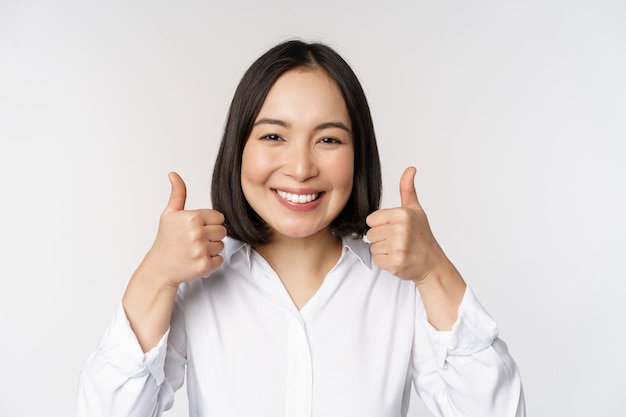 Rosto de mulher asiática alegre mostrando os polegares para cima e sorrindo satisfeito por algo que recomenda uma boa posição sobre fundo branco