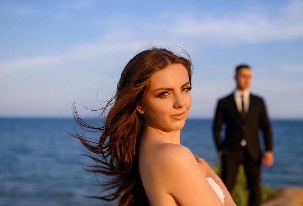 Rosto de retrato de noiva bonita no vestido de noiva com mangas abertas enquanto noivo elegante atrás de pé no fundo da paisagem do mar Sol brilhando no rosto atraente da mulher com maquiagem de casamento Natureza