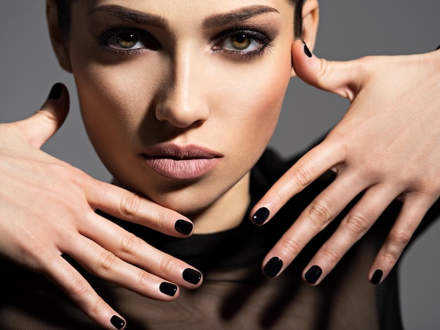 Rosto de uma linda garota com maquiagem fashion e unhas pretas posando sobre uma parede escura