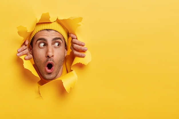 Rosto masculino surpreso pelo buraco do papel. jovem surpreso e emocional usando um capacete amarelo