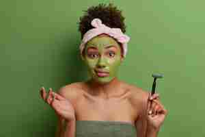 Foto grátis rotina matinal de cuidados com a pele. mulher inconsciente encolhe os ombros com hesitação, aplica máscara hidratante no rosto, segura a lâmina de barbear para fazer a barba, enrolada em uma toalha de banho, posa contra uma parede verde viva