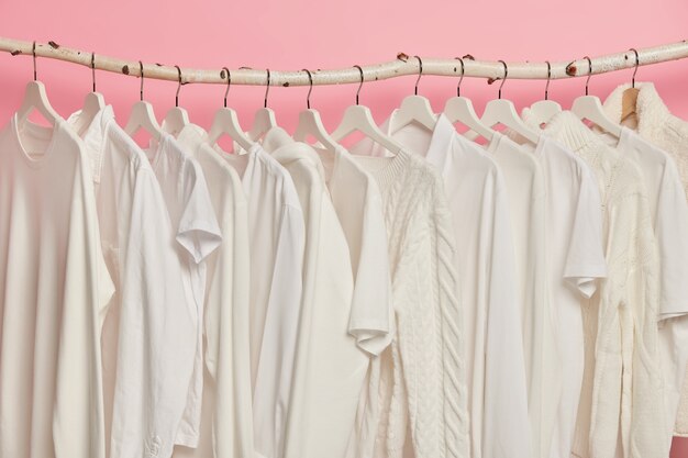 Roupas brancas sólidas penduradas em uma fileira em prateleiras de madeira contra um fundo rosa. Grande escolha para mulheres na loja de varejo.