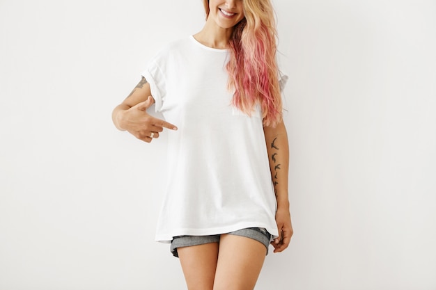 Roupas, design, estilo, moda e publicidade. Retrato de mulher irreconhecível hipster magro com destaques de cabelo rosa e braços tatuados, sorrindo, apontando o dedo para camiseta em branco