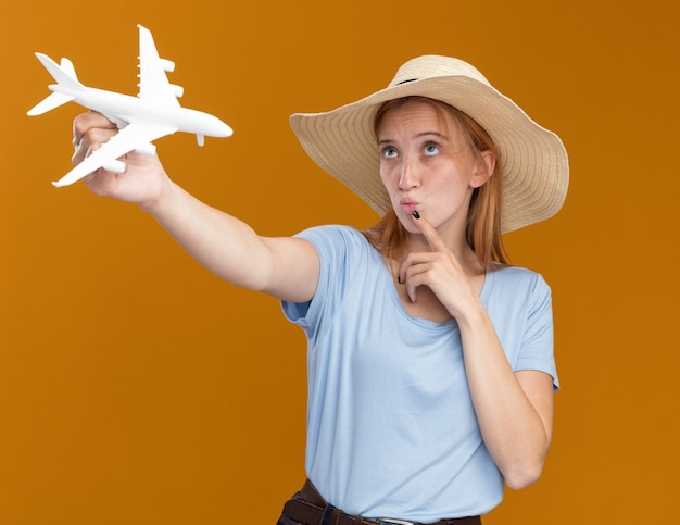 Ruiva jovem confusa e ruiva com sardas, usando um chapéu de praia, coloca o dedo no queixo e segura a modelo do avião, olhando para cima, isolada na parede laranja com espaço de cópia