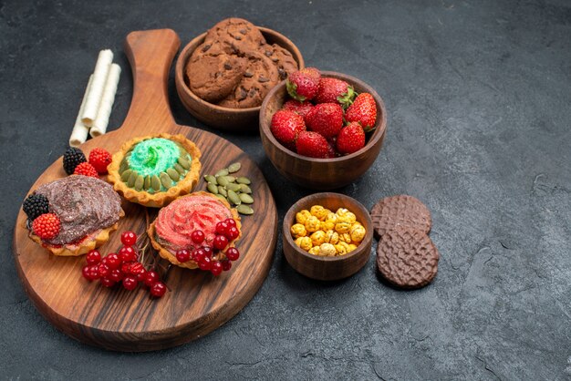 Saborosos bolos cremosos de vista frontal com biscoitos e frutas em fundo escuro