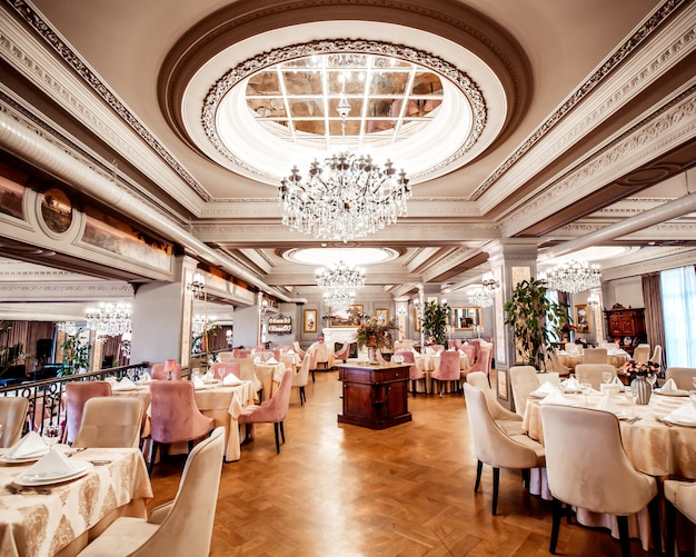 Sala de restaurante com mesas redondas e quadradas, algumas cadeiras e plantas