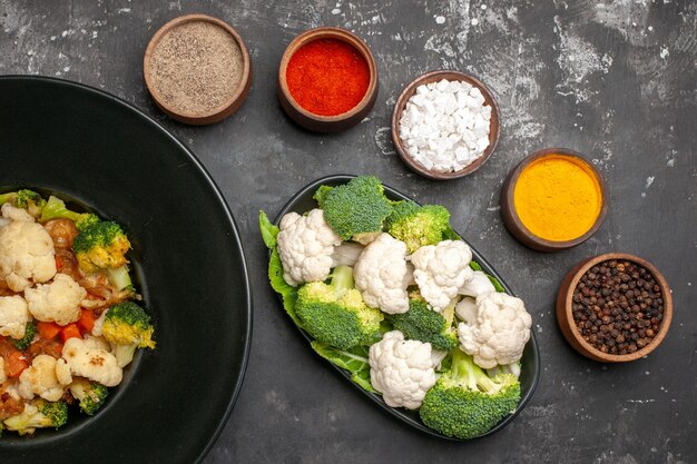 Salada de brócolis e couve-flor em uma placa oval preta com especiarias diferentes em pequenas tigelas brócolis e couve-flor crus em uma superfície escura