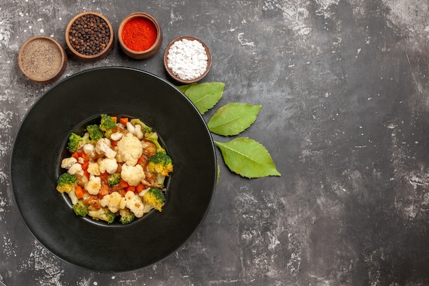 Salada de brócolis e couve-flor em uma tigela preta com especiarias diferentes em folhas de tigela na superfície escura com copa