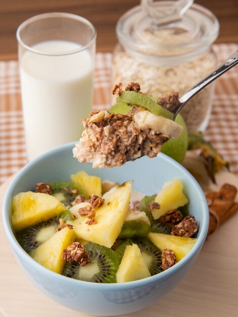 Salada de frutas de vista lateral em um prato kiwi abacaxi com granola em uma colher com um copo de leite