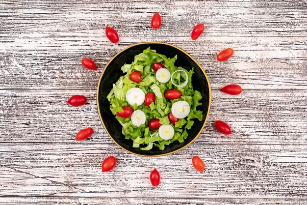 Salada de legumes com tomate cereja alface e cebola em uma tigela preta na superfície de madeira branca vista superior central