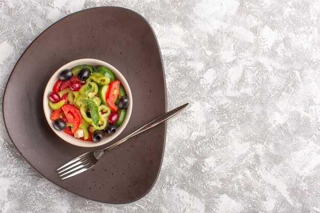 Salada de legumes fresca vista de cima com pepinos fatiados, tomates, azeitona e queijo branco dentro de um prato marrom na mesa rústica cinza