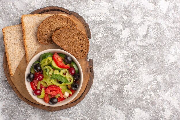 Salada de legumes fresca vista de cima com pepinos fatiados, tomates, azeitona e queijo branco dentro do prato com pão fatiado na mesa cinza salada de legumes