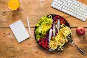 Foto grátis salada saudável com peixe cru, dispostas em placa; suco; maçã; teclado e bloco de notas; caneta na mesa de madeira