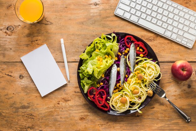 Salada saudável com peixe cru, dispostas em placa; suco; maçã; teclado e bloco de notas; caneta na mesa de madeira