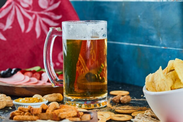 Salgadinhos variados, batatas fritas, um prato de salsichas e um copo de cerveja em superfície escura.