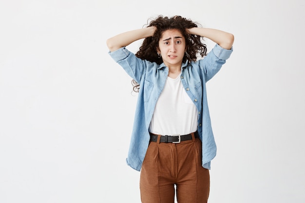 Foto grátis salientou intrigada jovem vestida casualmente com as mãos no cabelo escuro ondulado, sentindo tensão e estresse enquanto enfrentava problemas, não suporta pressão, cerra os dentes. linguagem corporal