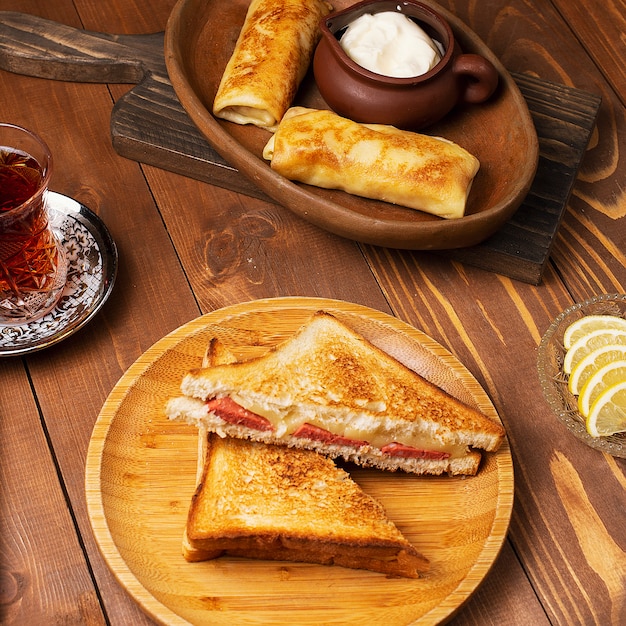 Sanduíches Club com salame, bacon e blinchik servido com iogurte na placa de madeira com chá