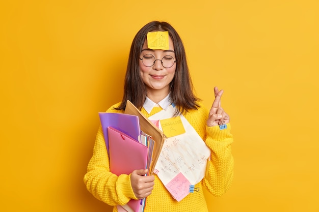 Foto grátis satisfeita, a aluna de asain acredita na boa sorte nas bancas de exames com os olhos fechados e os dedos cruzados, acredita que os sonhos se tornam realidade presa com papéis contém pastas.