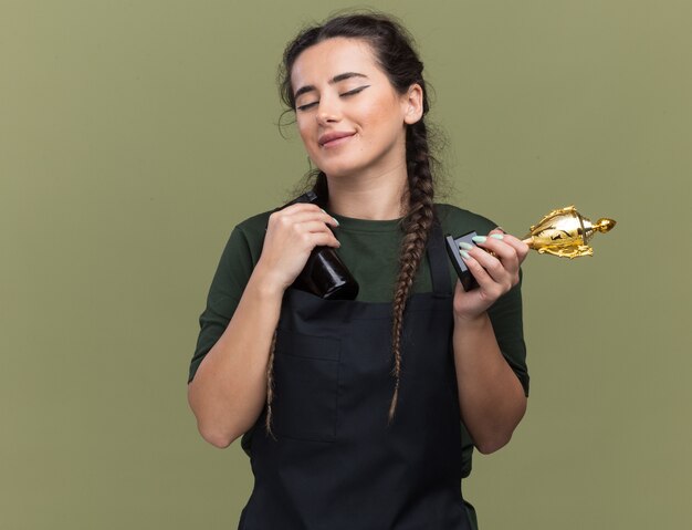 Satisfeita com os olhos fechados, jovem barbeira de uniforme segurando a taça do vencedor com uma tesoura de cabelo isolada na parede verde oliva