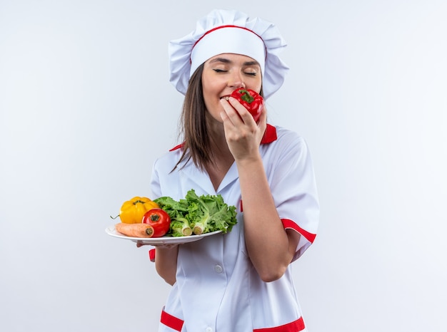 Satisfeita com os olhos fechados jovem cozinheira vestindo uniforme de chef segurando vegetais no prato cheirando pimenta isolada na parede branca