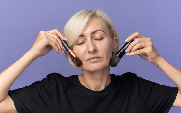 Satisfeita com os olhos fechados, uma jovem linda se senta à mesa com ferramentas de maquiagem, aplicando um blush em pó com um pincel de pó isolado na parede azul