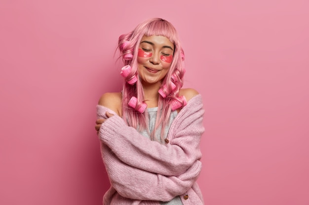 Foto grátis satisfeita e sorridente senhora encantadora fecha os olhos e se abraça, sente conforto no novo suéter de tricô, aplica protetores de beleza e rolinhos nos longos cabelos rosa